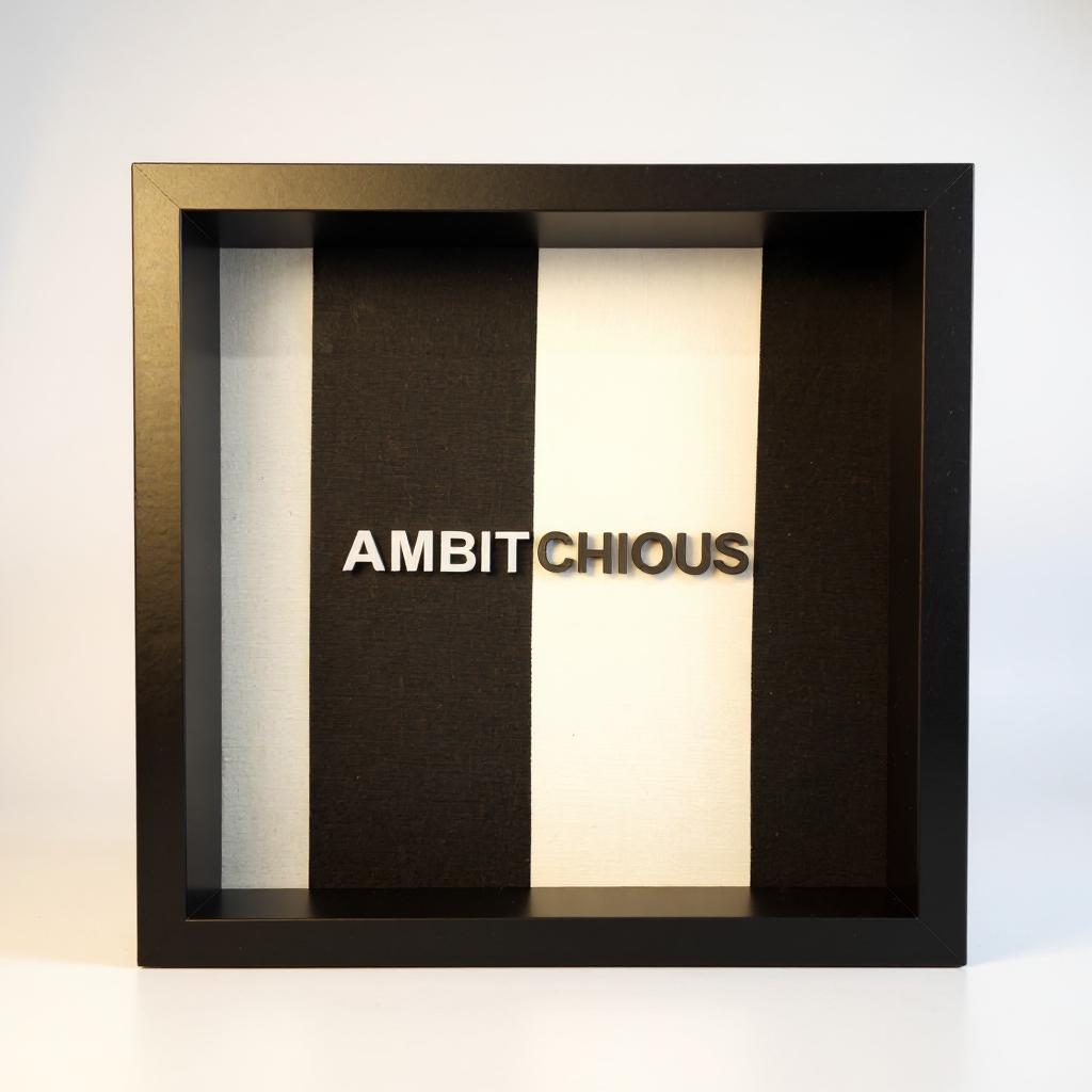 FACTIONARY™ Statement Frame „AMBITCHIOUS“ von OLDRYE®, schwarzer tiefer Rahmen mit erhabener 3D-Typografie auf schwarz-weiß gestreiftem Textilstoff, modernes Wortkunst-Design im Format 25x25 cm
